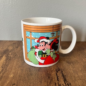Puede incluir: Taza de cer&aacute;mica blanca con Mickey y Minnie Mouse con atuendo navide&ntilde;o. Mickey lleva un gorro de Pap&aacute; Noel y un abrigo rojo, sosteniendo un saco verde. Minnie besa a Mickey bajo el mu&eacute;rdago. La taza tiene un asa y una escena invernal festiva.