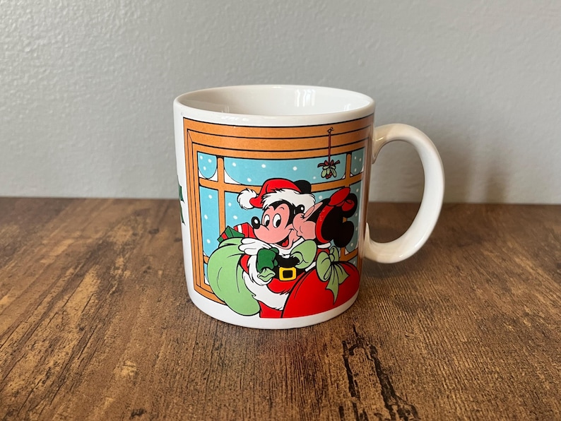 Puede incluir: Taza de cer&aacute;mica blanca con Mickey y Minnie Mouse con atuendo navide&ntilde;o. Mickey lleva un gorro de Pap&aacute; Noel y un abrigo rojo, sosteniendo un saco verde. Minnie besa a Mickey bajo el mu&eacute;rdago. La taza tiene un asa y una escena invernal festiva.