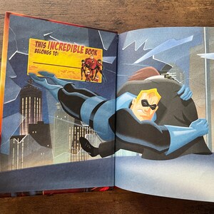 May include: An open hardcover book featuring an illustration of a superhero in a blue and black suit embracing a figure in a dark coat against a city skyline. A yellow rectangular label reads THIS INCREDIBLE BOOK BELONGS TO with the Incredibles family.