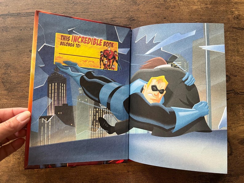 May include: An open hardcover book featuring an illustration of a superhero in a blue and black suit embracing a figure in a dark coat against a city skyline. A yellow rectangular label reads THIS INCREDIBLE BOOK BELONGS TO with the Incredibles family.