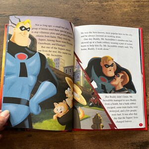 May include: An open children's storybook featuring illustrations of Mr. Incredible from The Incredibles. The left page shows Mr. Incredible standing tall, while the right page depicts him with Buddy. The text narrates the story of the superhero.