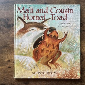 May include: A hardcover children's book titled Ma'ii and Cousin Horned Toad, A Traditional Navajo Story by Shonto Begay. The cover features a brown and white coyote illustration interacting with a brown horned toad against a textured background.