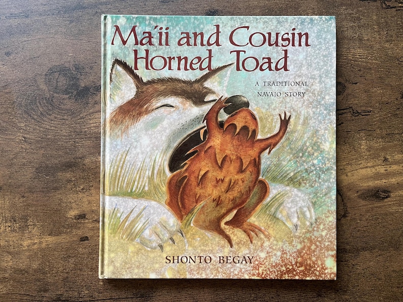 May include: A hardcover children's book titled Ma'ii and Cousin Horned Toad, A Traditional Navajo Story by Shonto Begay. The cover features a brown and white coyote illustration interacting with a brown horned toad against a textured background.