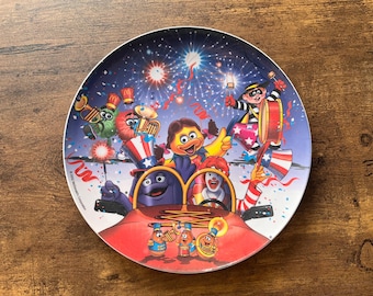 May include: A decorative plate featuring a festive scene with cartoon characters, fireworks, and patriotic imagery. The plate has a white rim and a colorful illustration of a parade with musical instruments and a car.