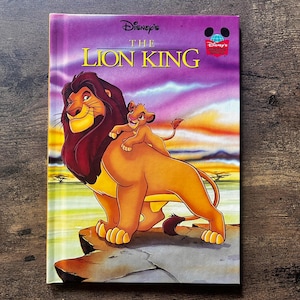 Puede incluir: Un libro de tapa dura titulado Disney's The Lion King con una colorida ilustración de Mufasa y Simba sobre una roca frente a un cielo al atardecer. La portada muestra el título en letras amarillas y un pequeño logotipo de Disney.