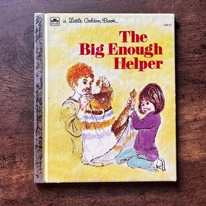 May include: A Little Golden Book titled The Big Enough Helper, featuring a yellow cover with an illustration of an adult and a child holding a striped blanket. The book has a silver spine and the number 208-51 printed in the top right corner.