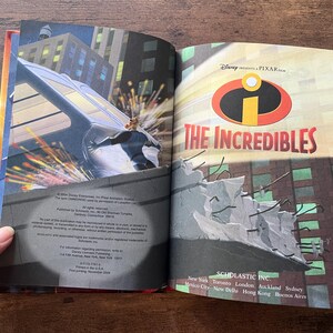 May include: An open hardcover book titled The Incredibles, featuring a Disney Pixar logo and a graphic illustration of a character pushing a vehicle on the left page, with copyright and publisher text printed on the bottom of the pages.