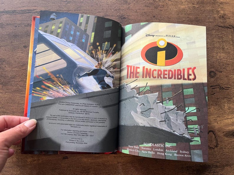 May include: An open hardcover book titled The Incredibles, featuring a Disney Pixar logo and a graphic illustration of a character pushing a vehicle on the left page, with copyright and publisher text printed on the bottom of the pages.