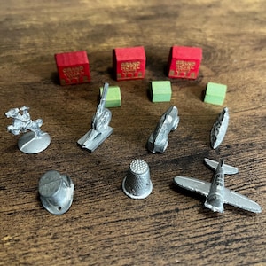 Puede incluir: Una colección de piezas del juego Monopoly, que incluyen un coche de carreras, un dedal y un avión. También hay piezas de hotel rojas con texto dorado y casas verdes. Las piezas son plateadas y están sobre una superficie de madera.