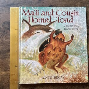 May include: A hardcover children's book titled Ma'ii and Cousin Horned Toad, A Traditional Navajo Story by Shonto Begay. The cover features a brown coyote and a horned toad. A yellow ruler shows the book height is approximately 10 inches.