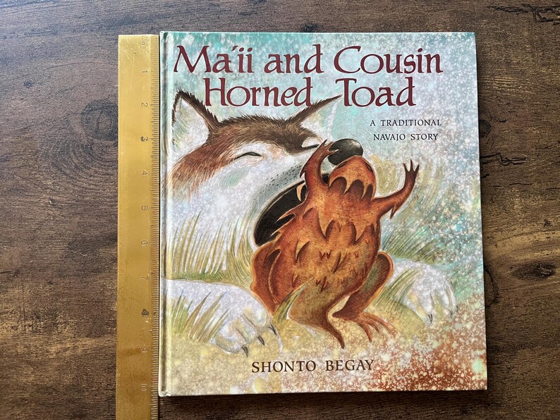 May include: A hardcover children's book titled Ma'ii and Cousin Horned Toad, A Traditional Navajo Story by Shonto Begay. The cover features a brown coyote and a horned toad. A yellow ruler shows the book height is approximately 10 inches.