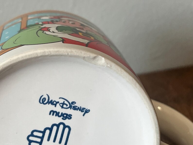 Puede incluir: Taza de cer&aacute;mica blanca con una colorida ilustraci&oacute;n de un personaje de dibujos animados. La taza tiene el texto "Walt Disney mugs" en azul. La taza es blanca con una peque&ntilde;a astilla en el borde.