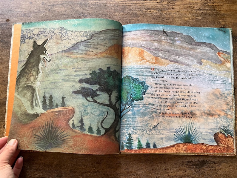 May include: An open children's book featuring a watercolor illustration of a coyote sitting on a rocky ledge overlooking a valley. The right page contains printed text describing the scene, with a mountain landscape and trees in the background.