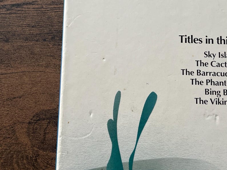 Op de afbeelding: Een witte boekomslag met een lijst van titels waaronder Sky Island, The Cactus, The Barracuda, The Phantom, Bing B en The Viking. Onder de tekst staat een gestileerde, blauwgroene grafische illustratie van waterplanten of zeewier.