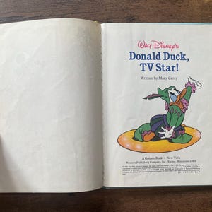 May include: An open children's book titled Walt Disney's Donald Duck, TV Star! written by Mary Carey. The right page features a colorful illustration of Donald Duck wearing a green and purple costume, sitting on a yellow oval, with text below.