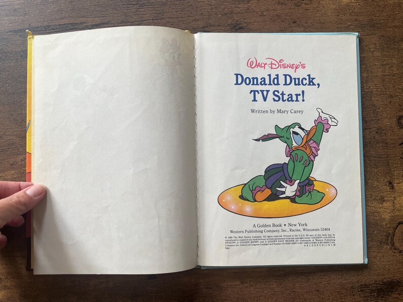 May include: An open children's book titled Walt Disney's Donald Duck, TV Star! written by Mary Carey. The right page features a colorful illustration of Donald Duck wearing a green and purple costume, sitting on a yellow oval, with text below.