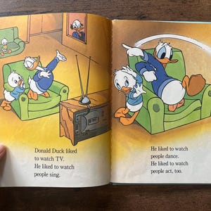 May include: An open children's book featuring colorful illustrations of Donald Duck and his nephews in a living room. The text reads: Donald Duck liked to watch TV. He liked to watch people sing. He liked to watch people dance. He liked to watch people act, too.