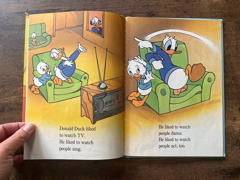 May include: An open children's book featuring colorful illustrations of Donald Duck and his nephews in a living room. The text reads: Donald Duck liked to watch TV. He liked to watch people sing. He liked to watch people dance. He liked to watch people act, too.