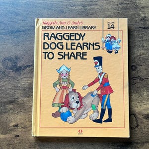 Puede incluir: Un libro infantil vintage titulado "Raggedy Dog Learns to Share" de la serie Grow-and-Learn Library. La portada presenta ilustraciones de Raggedy Ann, Andy, un perro y un soldado de juguete en un diseño colorido. Se muestra el volumen 14.