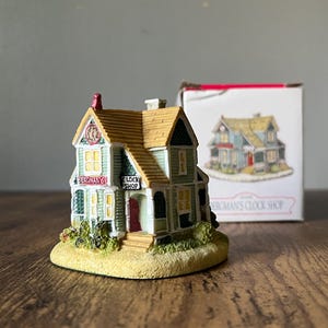 May include: A miniature house figurine with a tan roof, green and white facade, and the text "PERCHMAN'S CLOCK SHOP". The figurine sits on a small, grassy base. A box with a similar house illustration is in the background.