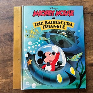Op de afbeelding: Een hardcover boek getiteld Disney's Mickey Mouse in The Barracuda Triangle, met Mickey Mouse in een blauwe onderzee&euml;r onder water. Een gele liniaal ligt langs de linkerkant en toont een hoogte van ongeveer 25 cm.
