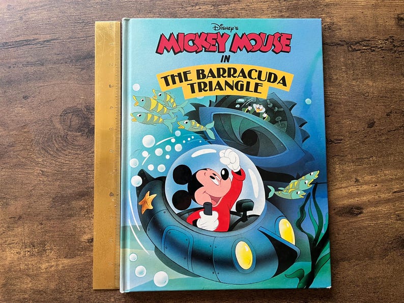 Op de afbeelding: Een hardcover boek getiteld Disney's Mickey Mouse in The Barracuda Triangle, met Mickey Mouse in een blauwe onderzee&euml;r onder water. Een gele liniaal ligt langs de linkerkant en toont een hoogte van ongeveer 25 cm.