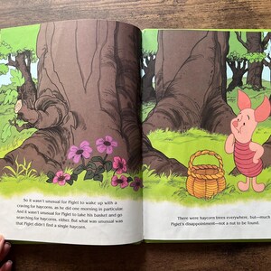 Puede incluir: Una p&aacute;gina de libro infantil abierta con una ilustraci&oacute;n colorida de Piglet de pie en un bosque junto a una cesta tejida. El texto dice: So it wasn't unusual for Piglet to wake up with a craving for haycorns, y There were haycorn trees everywhere, but&mdash;much to Piglet's disappointment&mdash;not a nut to be found.