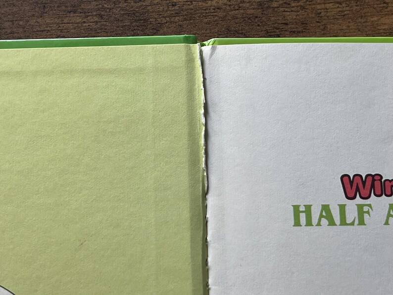 Puede incluir: Un primer plano de un libro abierto con una cubierta de color verde claro y p&aacute;ginas blancas. El lomo interior muestra signos de desgaste con bordes de papel deshilachados. En la p&aacute;gina derecha, el texto "Win" en rojo y "HALF A" en verde.