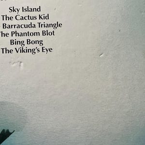 Op de afbeelding: Een witte boekomslag met een lijst van titels in zwarte tekst: Sky Island, The Cactus Kid, The Barracuda Triangle, The Phantom Blot, Bing Bong en The Viking's Eye. De onderrand toont een gedeeltelijk donkergroen grafisch element.