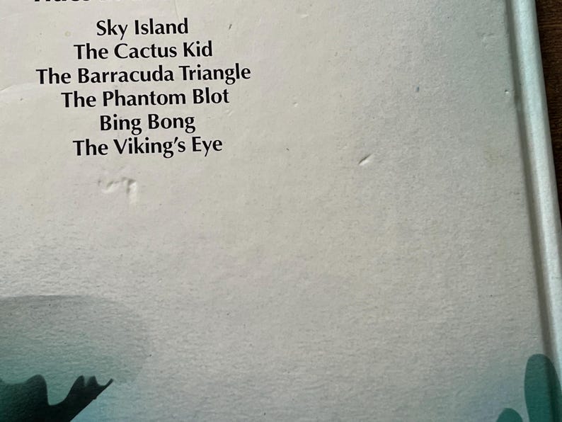Op de afbeelding: Een witte boekomslag met een lijst van titels in zwarte tekst: Sky Island, The Cactus Kid, The Barracuda Triangle, The Phantom Blot, Bing Bong en The Viking's Eye. De onderrand toont een gedeeltelijk donkergroen grafisch element.
