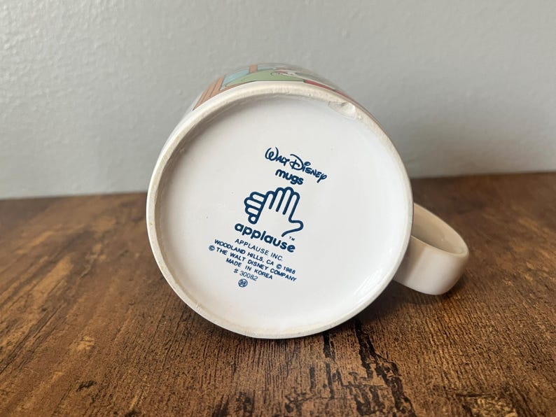 Puede incluir: Taza de cer&aacute;mica blanca de Disney con asa. La base de la taza presenta el texto "Walt Disney Mugs" y "applause" con un logotipo de mano. La taza est&aacute; hecha en Corea.