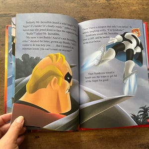 May include: An open children's storybook featuring a scene from The Incredibles. The left page shows Mr. Incredible in profile, while the right page depicts the character Syndrome flying with rocket boots above water, accompanied by printed narrative text.