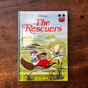 Puede incluir: Un libro de tapa dura titulado Disney's The Rescuers con una colorida ilustración de dos ratones en un bote de hoja con una libélula. La portada muestra el título en texto rojo, un logotipo de Disney y un paisaje verde con agua.