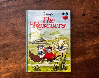 May include: A hardcover book titled Disney's The Rescuers featuring a colorful illustration of two mice in a leaf boat with a dragonfly. The cover displays the title in red text, a Disney logo, and a green landscape with water and reeds.