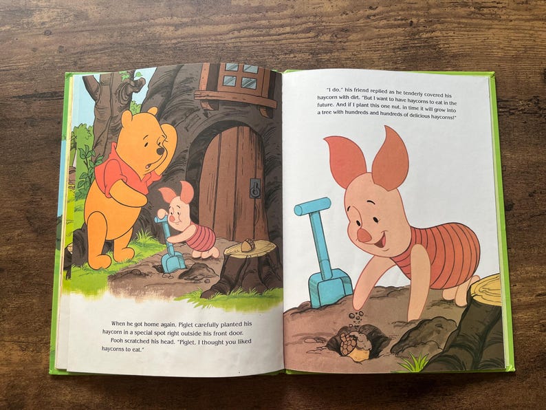Puede incluir: Un libro de cuentos infantil abierto con Winnie the Pooh y Piglet. La p&aacute;gina izquierda muestra a los personajes cerca de una casa en el &aacute;rbol, mientras que la derecha muestra a Piglet plantando una bellota con una pala azul. Texto incluido.