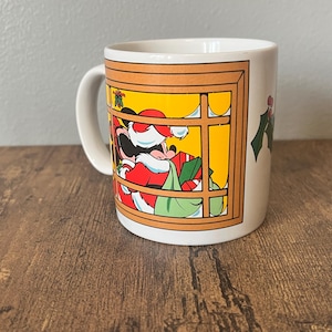 Puede incluir: Una taza de cer&aacute;mica blanca con un dise&ntilde;o navide&ntilde;o. La taza muestra un personaje de dibujos animados vestido de Pap&aacute; Noel, enmarcado por una ventana. El personaje sostiene un saco de regalos. Ramas de acebo adornan el lateral.