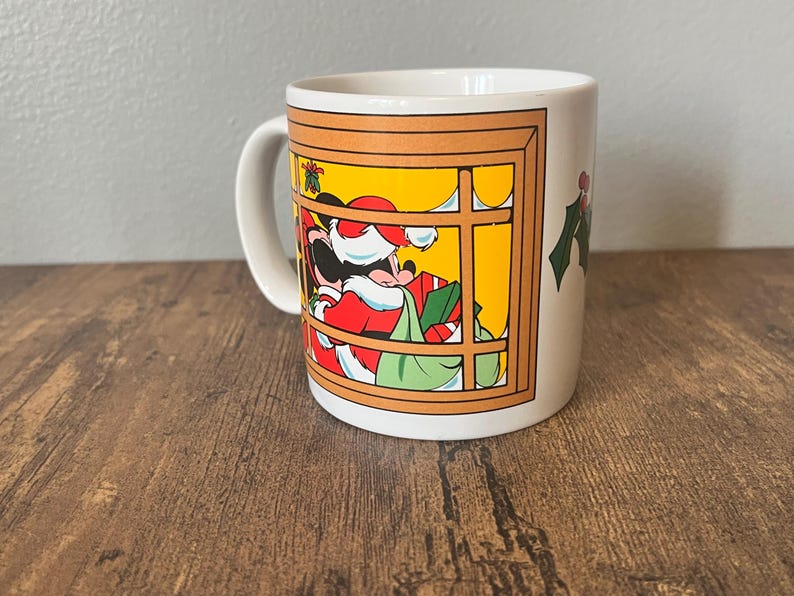 Puede incluir: Una taza de cer&aacute;mica blanca con un dise&ntilde;o navide&ntilde;o. La taza muestra un personaje de dibujos animados vestido de Pap&aacute; Noel, enmarcado por una ventana. El personaje sostiene un saco de regalos. Ramas de acebo adornan el lateral.