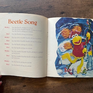 May include: An open children's book page featuring the text Beetle Song and lyrics alternating between Mokey and Red. The right page displays a colorful illustration of a yellow puppet character with orange hair wearing a red sweater.