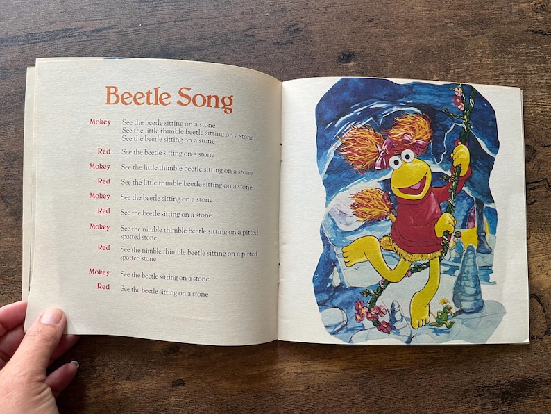 May include: An open children's book page featuring the text Beetle Song and lyrics alternating between Mokey and Red. The right page displays a colorful illustration of a yellow puppet character with orange hair wearing a red sweater.