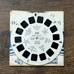 May include: Vintage View-Master reel featuring scenes from Sun Valley, Idaho, including Main Terrace, Ice Rink, and Dollar Mountain. The reel is white with black rectangular viewing windows and text. The reel is on a blue and white cardboard backing.