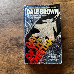 May include: A hardcover book titled "Day of the Cheetah" by Dale Brown. The cover features a silver jet graphic over a black background with red and yellow text. The book is a New York Times bestseller.