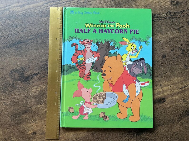 Puede incluir: Un libro de tapa dura Big Golden Book titulado Walt Disney's Winnie the Pooh Half a Haycorn Pie, con ilustraciones de Pooh, Piglet, Tigger, Rabbit y Eeyore. Una regla dorada de 30 cm est&aacute; colocada verticalmente a lo largo del lado izquierdo.