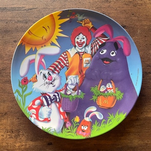 May include: A decorative plate featuring Ronald McDonald, Grimace, and Easter Bunny characters. The plate has a blue background with a smiling sun and green grass. The characters are holding Easter baskets.