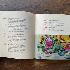 May include: An open children's book with two pages of printed text and a colorful illustration. The left page features a poem with red character names. The right page contains more text and a drawing of a pink puppet character writing in a book.