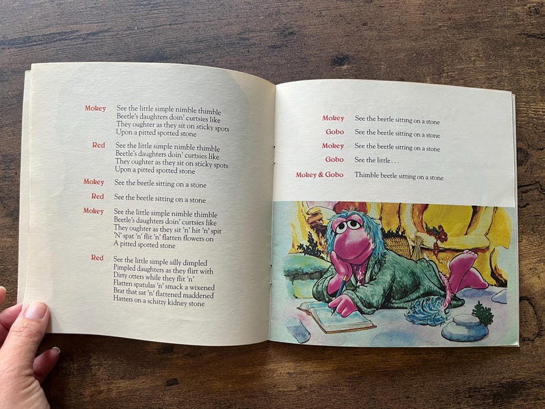 May include: An open children's book with two pages of printed text and a colorful illustration. The left page features a poem with red character names. The right page contains more text and a drawing of a pink puppet character writing in a book.