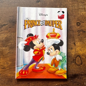 Puede incluir: Un libro de tapa dura titulado Disney's The Prince and the Pauper con una ilustración de dos personajes de Mickey Mouse en la portada. Un personaje viste un traje real rojo y dorado y coloca una corona dorada sobre la cabeza del otro.
