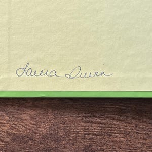 Puede incluir: Una superficie de papel rectangular de color amarillo claro con un borde verde brillante. El papel presenta la firma manuscrita Laura Durn en tinta azul oscuro centrada cerca del borde inferior de la secci&oacute;n amarilla.