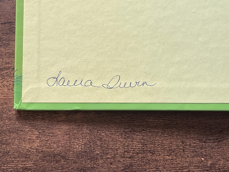 Puede incluir: Una superficie de papel rectangular de color amarillo claro con un borde verde brillante. El papel presenta la firma manuscrita Laura Durn en tinta azul oscuro centrada cerca del borde inferior de la secci&oacute;n amarilla.
