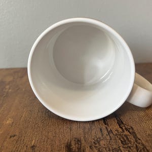Puede incluir: Una taza de cer&aacute;mica blanca con un asa redondeada. El interior de la taza es blanco y liso. La taza est&aacute; sobre una superficie de madera marr&oacute;n.
