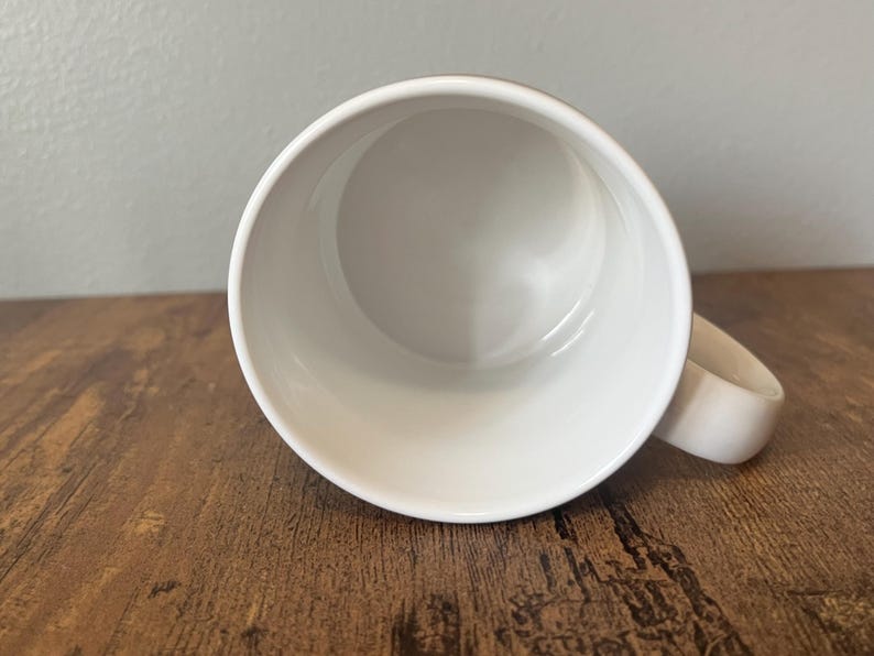 Puede incluir: Una taza de cer&aacute;mica blanca con un asa redondeada. El interior de la taza es blanco y liso. La taza est&aacute; sobre una superficie de madera marr&oacute;n.